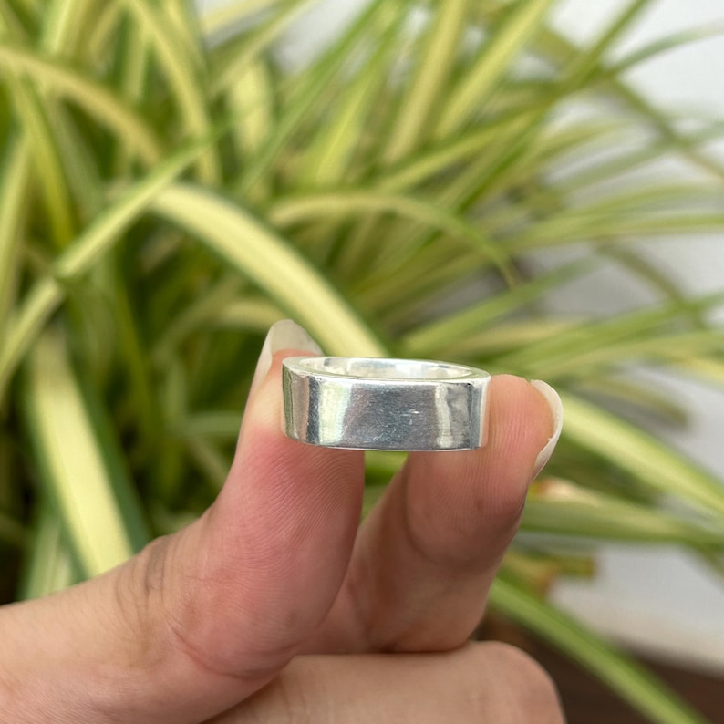 May include: A silver-colored, rectangular ring. The ring is held between fingers against a backdrop of green and yellow foliage. The ring's design is simple and modern, with a flat top and sides.