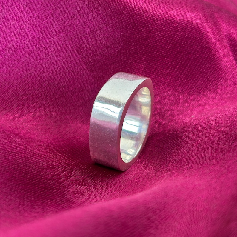 May include: A silver ring with a flat, rectangular design. The ring has a smooth, polished surface and a rounded interior. The ring is set against a deep pink background.
