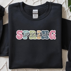 Pode incluir: Camisola preta com a palavra "SPRING BLOOM" em letras coloridas e decorativas. As letras são contornadas em branco e preenchidas com cores pastel e desenhos florais. A camisola está dobrada sobre uma superfície clara.