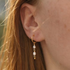 Peut inclure: Boucle d'oreille dorée avec un design pendant. La boucle d'oreille comporte une perle blanche, une petite perle verte et une perle dorée. La boucle d'oreille est portée à l'oreille d'une personne, avec ses cheveux visibles en arrière-plan.