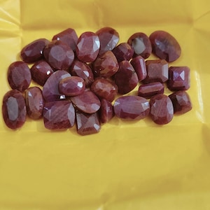 May include: A collection of numerous loose, faceted, deep red rubies in various shapes including oval, round, and rectangular cuts. These gemstones exhibit a polished, reflective surface with visible internal inclusions, arranged in a cluster.