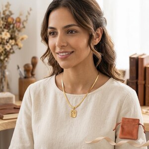 Puede incluir: Un collar de cadena de espiga en tono dorado con un colgante rectangular que presenta un diseño de rostro en arte lineal abstracto. El collar se acompaña de una pequeña bolsa de terciopelo marrón con cordón.