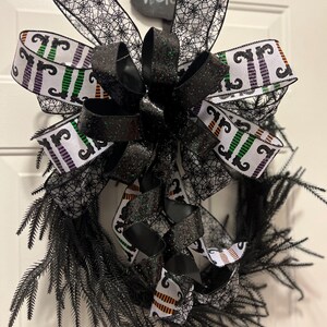 May include: A black Halloween wreath with a large bow featuring spiderweb and striped ribbon. The wreath has black, feathery branches and is decorated with a chalkboard sign that says "Welcome".
