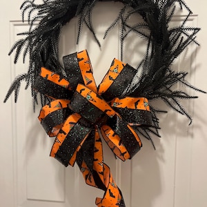 Può includere: Una ghirlanda nera di Halloween con un grande fiocco. La ghirlanda è realizzata in materiale scuro e appuntito. Il fiocco presenta nastri arancioni e neri con un motivo a pipistrelli e cappelli da strega e accenti di glitter neri.