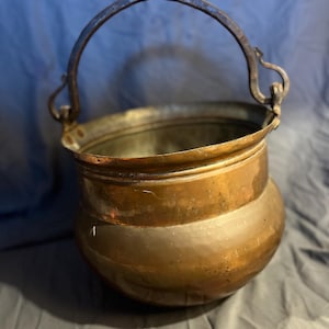 May include: A copper cauldron with a curved handle. The pot has a rounded body and a wide opening. The handle is made of a darker metal and is attached to the sides of the cauldron. The cauldron has a rustic, antique appearance.