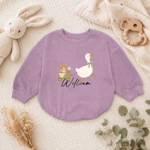 May include: A lavender long-sleeve sweatshirt featuring a graphic of a white goose pulling a small green wagon with a brown teddy bear inside. The name William is printed in black cursive script below the illustration. The garment has a ribbed crew neckline.