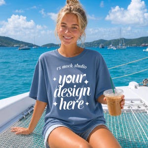 Può includere: Una t-shirt blu a maniche corte con il testo bianco rs mock studio your design here e accenti a forma di stella. La maglia presenta un girocollo e una vestibilità rilassata, mostrata su uno sfondo di acqua di mare blu e colline lontane.