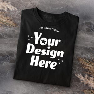 Op de afbeelding: Zwart T-shirt met de tekst "Your Design Here" in een vet, wit lettertype, met een ster die de "O" in "Your" vervangt. De tekst "RS MOCK STUDIO" staat boven de hoofdtekst. Het shirt is op een grijs oppervlak gevouwen.