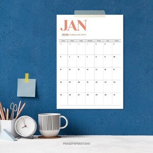 May include: A white 2026 calendar page for January, taped to a blue wall. The word "JAN" is in coral, with "Walking with JESUS" below. A clock, mug, and stationery are on a white surface.