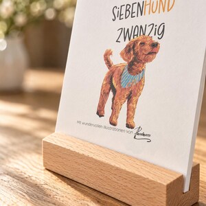 May include: A white rectangular card featuring a brown dog illustration wearing a blue striped bandana, with the text ZWEITAUSEND SIEBENHUND ZWANZIG and Mit wundervollen Illustrationen von Pinselmessi, held upright in a light-colored wooden block.