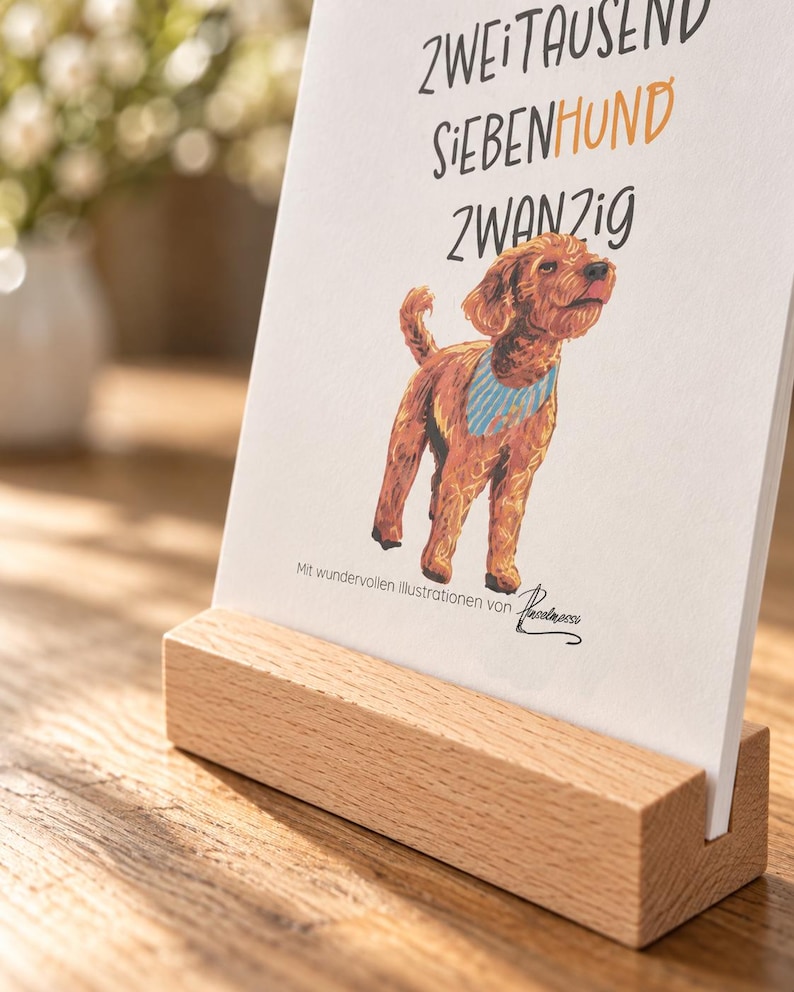 May include: A white rectangular card featuring a brown dog illustration wearing a blue striped bandana, with the text ZWEITAUSEND SIEBENHUND ZWANZIG and Mit wundervollen Illustrationen von Pinselmessi, held upright in a light-colored wooden block.