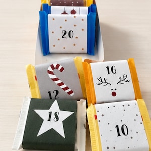 May include: A collection of small, rectangular, individually wrapped chocolate bars featuring festive designs and numbers. The wrappers display various holiday motifs including a candy cane, reindeer antlers, stars, and dots, with numbers 10, 12, 16, 18, and 20.