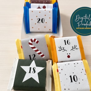 May include: A collection of small, rectangular chocolate bars wrapped in festive paper with various holiday designs, including numbers 10, 12, 16, 18, and 20, candy canes, stars, and reindeer antlers, displayed in a white cardboard tray.