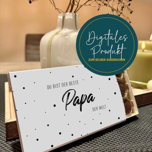 Pode incluir: Uma caixa de presente branca com bolinhas pretas e as palavras "DU BIST DER BESTE Papa DER WELT" em escrita preta. Um círculo verde-azulado com as palavras "Digitales Produkt" está no fundo. Um vaso e velas também estão no fundo.