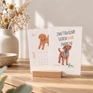 May include: A desk calendar featuring a light wood block stand holding two white cards. The front card displays a brown poodle illustration, the text ZWEITAUSEND SIEBENHUND ZWANZIG, and a signature. The back card shows a January calendar page.