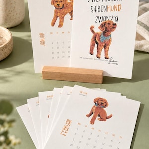 May include: A set of white paper calendar pages featuring illustrations of brown dogs. The top page displays the text ZWEITAUSEND SIEBENHUND ZWANZIG and a dog wearing a blue bandana. Below, a page shows the month FEBRUAR with a dog illustration.