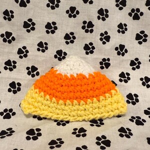 Könnte beinhalten: Ein gehäkeltes Candy Corn in Gelb, Orange und Weiß. Das Candy Corn liegt auf einem weißen Hintergrund mit schwarzen Pfotenabdrücken. Das Candy Corn ist ein Halloween-Artikel.