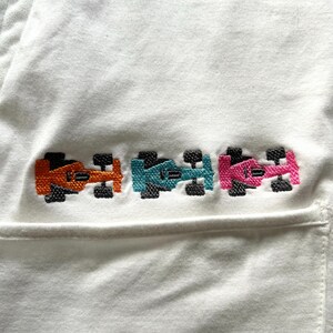 May include: White fabric with an embroidered design of three race cars in orange, teal, and pink. The cars have black wheels and are arranged horizontally. The fabric appears to be a pocket or part of a garment.