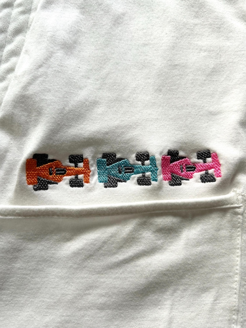 May include: White fabric with an embroidered design of three race cars in orange, teal, and pink. The cars have black wheels and are arranged horizontally. The fabric appears to be a pocket or part of a garment.