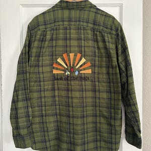 May include: An olive green and black plaid long-sleeve button-down shirt featuring an embroidered design on the back. The embroidery depicts a sunburst in shades of orange and yellow, small insect figures, and the text "last of the bugs".