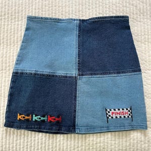 May include: A denim skirt with a patchwork design in shades of blue. The skirt features embroidered race cars and a checkered flag with the word "FINISH".