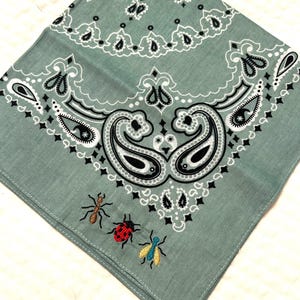 May include: A sage green bandana with a paisley design and embroidered insects. The bandana has a detailed white, black, and grey paisley pattern along the edges and a central design. Three embroidered insects, an ant, a ladybug, and a fly, are in the corner.