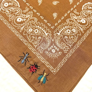 May include: A brown bandana with a white paisley pattern and embroidered insects. The insects include a blue ant, a red ladybug, and a blue and yellow fly. The bandana is square and made of a soft fabric.