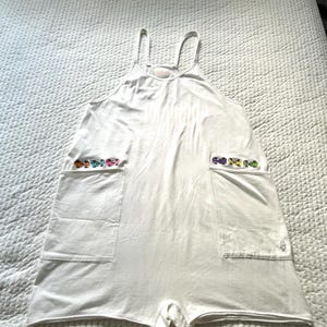 May include: White romper with spaghetti straps and two large front pockets. Each pocket is decorated with a row of colourful, embroidered toy cars. The romper is made of a soft, lightweight fabric.