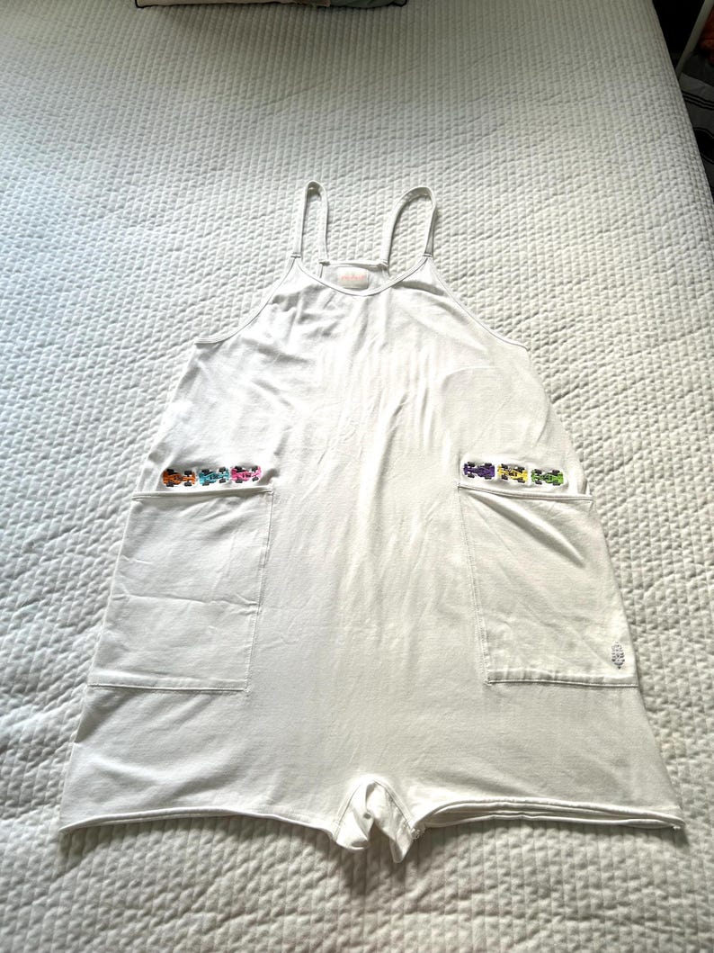 May include: White romper with spaghetti straps and two large front pockets. Each pocket is decorated with a row of colorful, embroidered toy cars. The romper is made of a soft, lightweight fabric.