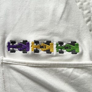 May include: White garment featuring an embroidered design of three pixelated race cars in purple, yellow, and green. The cars have black wheels and are aligned horizontally. The garment has a pocket.