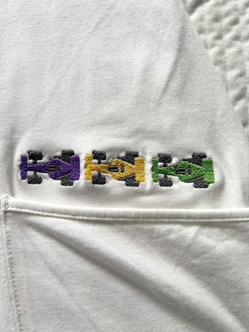 May include: White garment featuring an embroidered design of three pixelated race cars in purple, yellow, and green. The cars have black wheels and are aligned horizontally. The garment has a pocket.