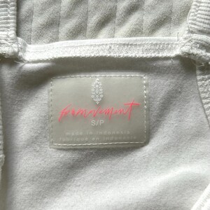 May include: Close-up of a white ribbed fabric garment. A rectangular label with the words "movement" in pink script is visible. The label also includes the size "S/P" and the text "made in Indonesia".
