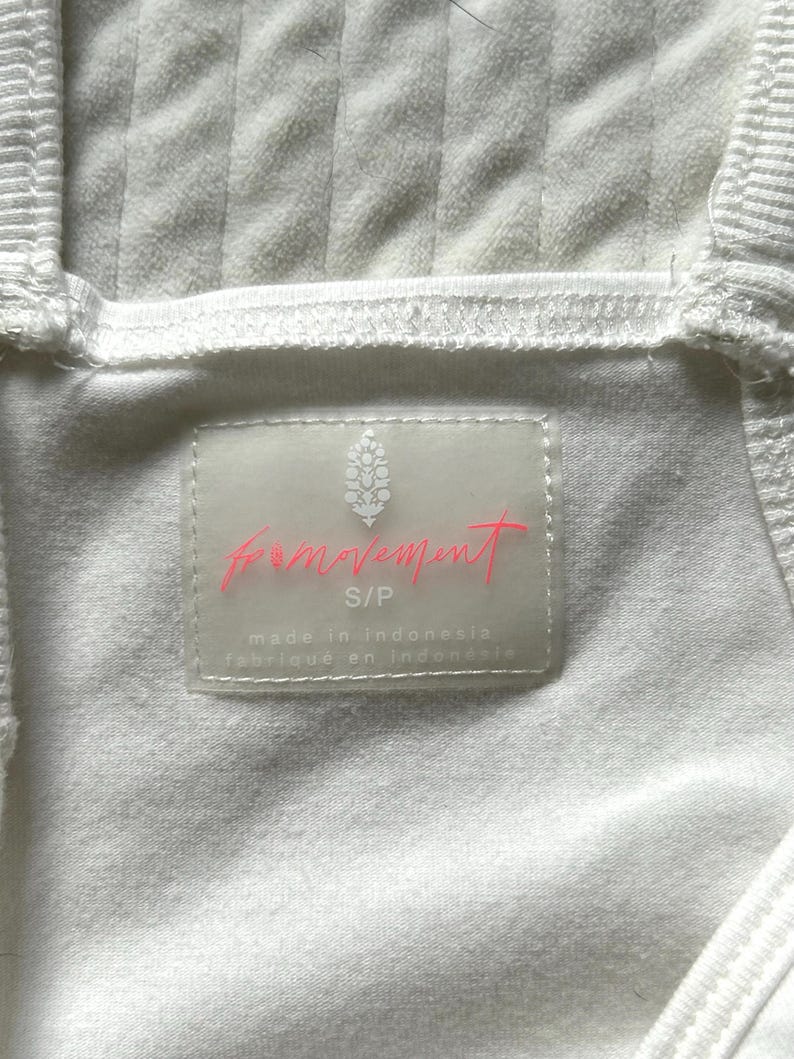 May include: Close-up of a white ribbed fabric garment. A rectangular label with the words "movement" in pink script is visible. The label also includes the size "S/P" and the text "made in Indonesia".