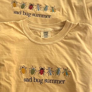 May include: A light yellow t-shirt featuring an embroidered row of six colourful insects above the black text "sad bug summer". The shirt has a crew neckline and a Comfort Colors brand label visible on the inside of the collar area.