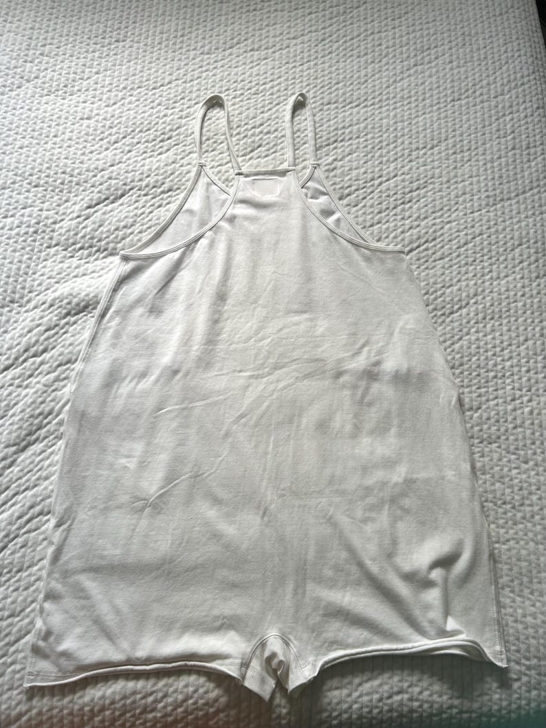 May include: A white romper with thin spaghetti straps and a scoop neckline. The one-piece garment has a relaxed fit and short legs. The fabric appears to be a soft, lightweight material, suitable for warm weather.