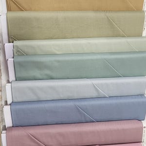 May include: A display of fabric rolls in various pastel colors, including pink, beige, and blue. The fabric is neatly folded and presented in a stacked arrangement, showcasing a range of soft hues.