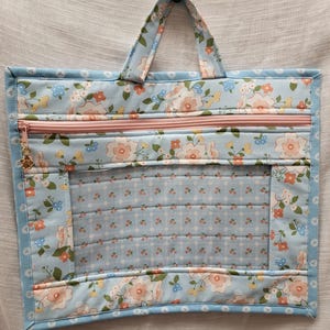 May include: A light blue quilted tote bag with a floral pattern in pink, white, and blue. The bag has a pink zipper, a clear window, and a matching handle. The bag is approximately 12 inches wide and 10 inches tall.