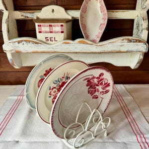 May include: A collection of vintage-style plates with red floral designs, displayed in a white metal plate holder. A white shelf with a salt container labeled "SEL" and a decorative dish are in the background. The plates rest on a white tablecloth with red stripes.