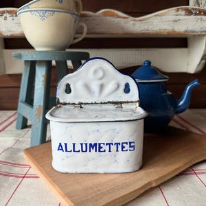 Könnte beinhalten: Eine weiß-blaue Emaille-Streichholzschachtel mit dem Wort "ALLUMETTES" in blauen Buchstaben. Die Streichholzschachtel hat einen Klappdeckel und steht auf einem Holzbrett. Eine blaue Teekanne und zwei cremefarbene Tassen sind im Hintergrund.