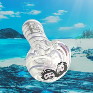 May include: Clear glass pipe with a swirling, striped design. The bowl features a black and white illustration of two faces. The background is a beach scene with blue water and sky.