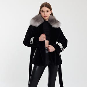 May include: A black faux fur coat with a grey fur collar and decorative cuffs. The coat has a matching belt and is worn with black leggings and ankle boots. The overall style is chic and fashionable.