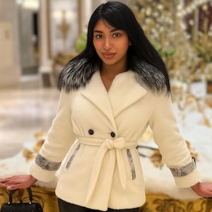 May include: Cream-colored coat with a faux fur collar and a matching belt. The coat features a double-breasted design with black buttons and snake-print detailing on the cuffs and pockets. A black handbag with a sparkly finish is held in the hand.