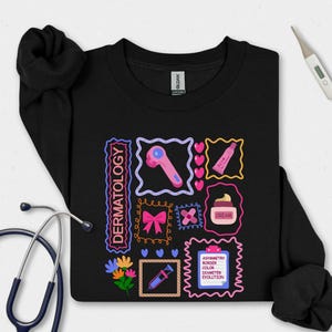 May include: Black sweatshirt with a colorful dermatology-themed graphic. The design includes illustrations of skincare tools, a bow, hearts, and the word "DERMATOLOGY". A stethoscope and thermometer are also visible.
