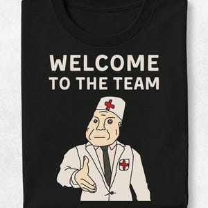 May include: A black t-shirt featuring the white text "WELCOME TO THE TEAM" above a graphic of a medical professional in a white coat, tie, and cap with a red cross symbol, extending a hand forward in a gesture of greeting or introduction.
