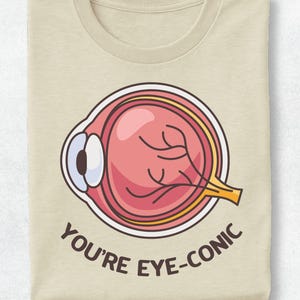 Könnte beinhalten: Ein beiges T-Shirt mit einer grafischen Darstellung eines anatomischen menschlichen Auges in Rosa-, Weiß- und Gelbtönen. Unter dem Auge ist der Text "YOU'RE EYE-CONIC" in dunkelbraunen Großbuchstaben mittig auf der Vorderseite aufgedruckt.