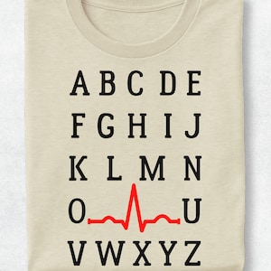 May include: A beige t-shirt featuring a black printed alphabet from A to Z arranged in five rows. The letter P is replaced by a red EKG heart rhythm line that spans the space where the letter would be, creating a visual medical heartbeat graphic.