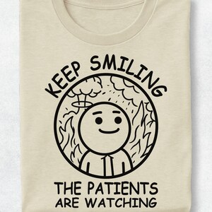May include: A beige t-shirt featuring a black circular graphic of a smiling character standing amidst flames and a mushroom cloud. The text reads KEEP SMILING above the circle and THE PATIENTS ARE WATCHING below the graphic in a bold font.
