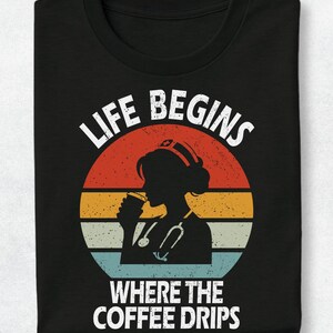 May include: A black t-shirt featuring a graphic print of a nurse silhouette holding a coffee cup with a stethoscope around the neck. The design includes a retro-style sunset background in red, orange, and blue, with the text LIFE BEGINS WHERE THE COFFEE DRIPS.