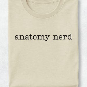 May include: A beige crew neck t-shirt featuring the text anatomy nerd printed in a simple black typewriter-style font across the center of the chest. The garment has short sleeves and a standard fit, presented in a flat, folded orientation.