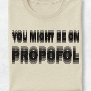 May include: A beige crew neck t-shirt featuring the black text "YOU MIGHT BE ON PROPOFOL" printed on the front. The lettering has a layered, drop-shadow effect that creates a blurred, motion-like appearance across the chest of the garment.
